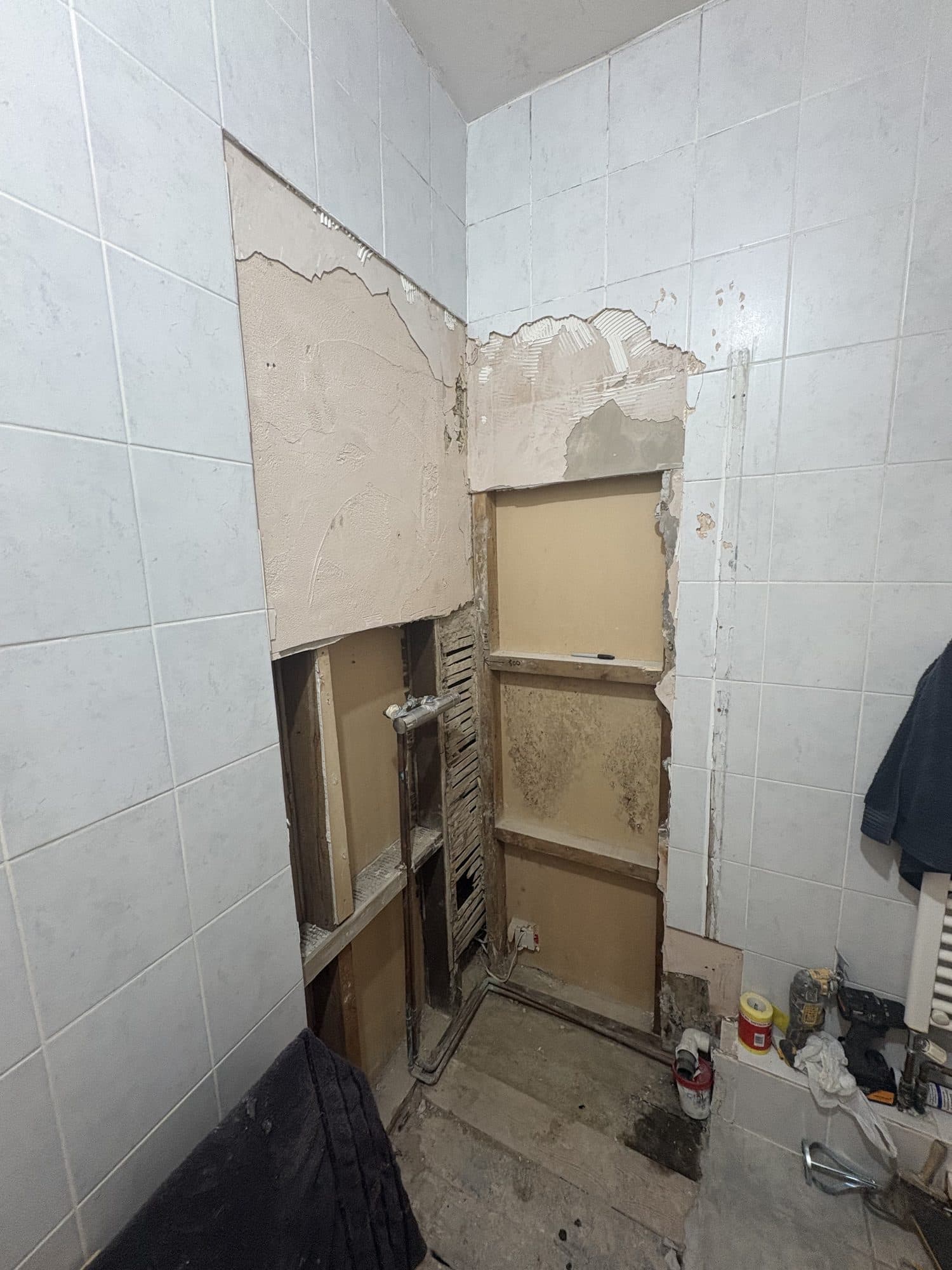 Bathroom wall with a section of tiles cut out, exposing the boards and pipework behind it during the work