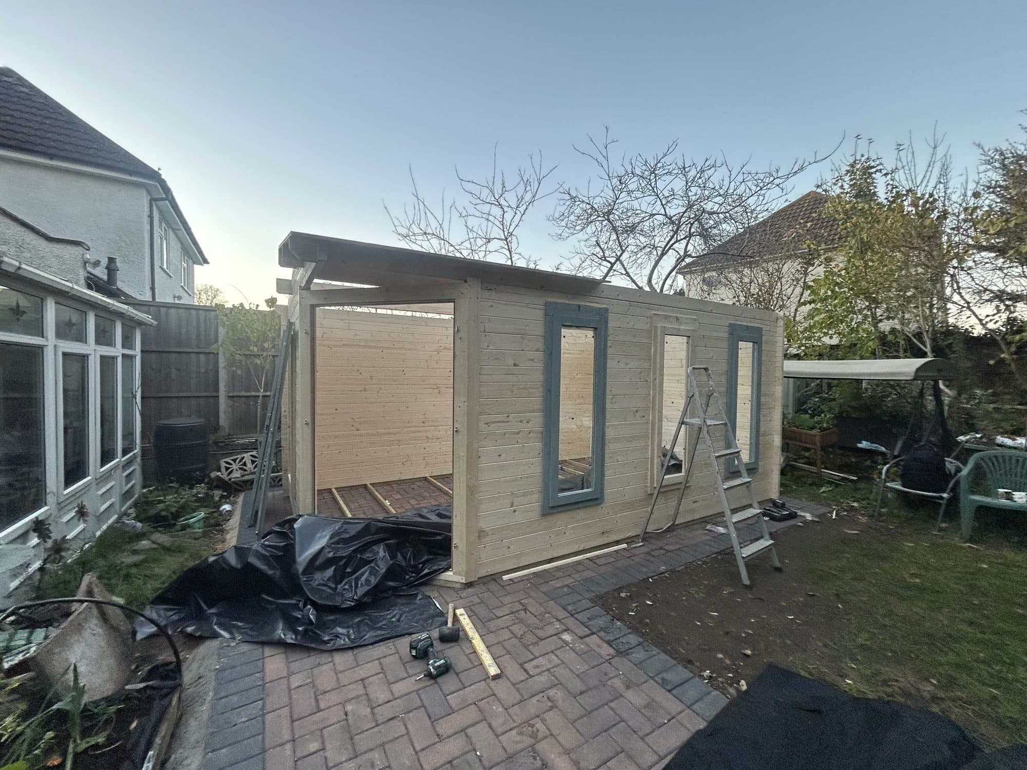 Timber summer house frame under construction on a paved base in a Uxbridge garden, with a stepladder and power tools beside it