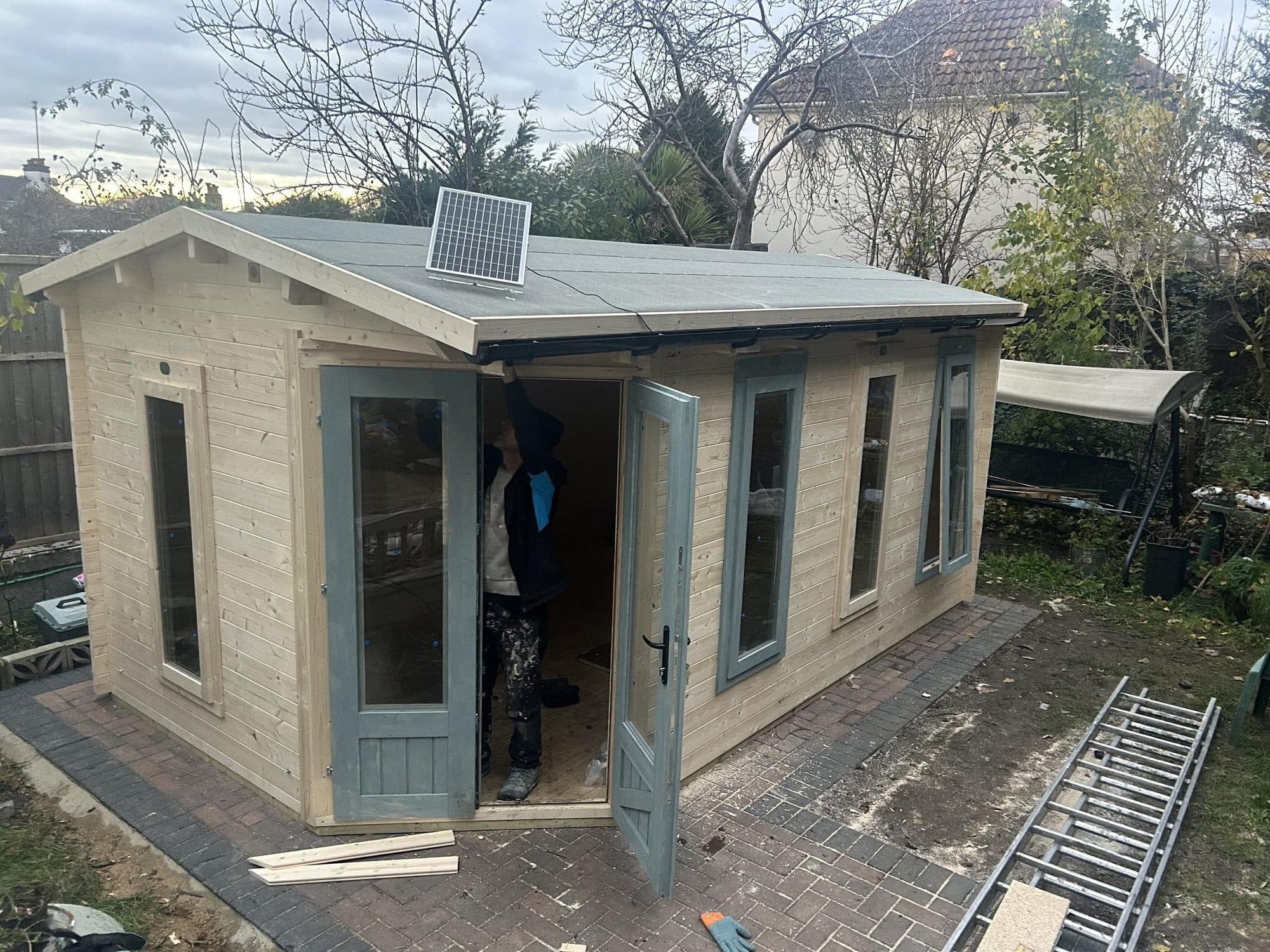 Completed timber summer house with grey-green double doors and windows, standing on a paved base